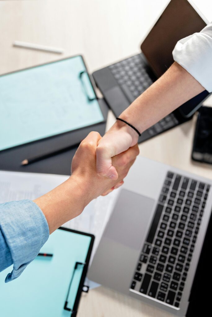 Two people shaking hands over laptops and documents in an office, symbolizing a business deal.